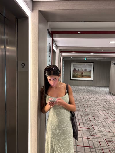 A young woman with long dark hair stands in a hotel hallway on the 9th floor of a building located in San Miguel de Cozumel, Mexico. She is leaning with her right shoulder against a light-colored vertical wall panel that features a thin illuminated strip light on its left edge. She wears a light green spaghetti-strap dress with a small white floral pattern, a gold-colored choker necklace, and dark sunglasses perched on her head. A dark shoulder bag hangs from her left shoulder. Her gaze is directed downwards as she intently operates a white smartphone with both hands. To her immediate left is a closed stainless steel elevator door, next to which a gray plaque displays the number "9". The hallway extends into the background, carpeted with a complex geometric pattern of gray, white, and red lines. The ceiling is white with recessed lighting, bordered by a distinct red horizontal stripe along the upper wall trim. On the far wall in the background, a large framed landscape photograph depicts a desert scene with prominent rock formations and a lone figure. Further down the hallway on the right, a sign with an arrow pointing right is partially visible.