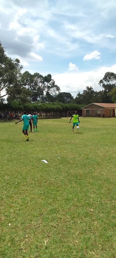 The image depicts a soccer field in Lubao, Kenya, located at coordinates 0.384 latitude and 34.801 longitude. Four young men are playing soccer on a green field, under a partly cloudy sky. The players are wearing green and yellow jerseys. There are trees and a house in the background. The players are in the middle of a game, with one player dribbling the ball while the other three are running around him. The field is green and well-kept. The players are in a relaxed and playful mood, enjoying the game.