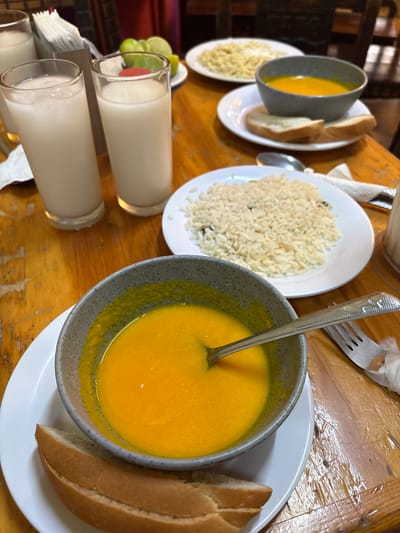 The image depicts a wooden table laden with food and drinks, likely in a casual dining setting. In the foreground, a grey bowl filled with bright orange soup is placed on a white plate, accompanied by two slices of bread. A spoon rests in the soup, and a fork and knife are visible to the right. Behind this, two tall glasses filled with a milky-white beverage, possibly horchata or a similar drink, stand next to a dispenser of white paper napkins. Further back, a plate of white rice and another plate with pasta and a small bowl of orange soup are partially visible. Lime wedges are arranged in a small dish near the back of the table. The overall impression is of a meal in progress. The wooden table has a visible grain and some wear, suggesting it might be in a restaurant or home. The background is blurred but hints at a dimly lit interior with chairs and other tables. Based on the presence of horchata, lime, and the general culinary style, this scene could be set in Cuernavaca, Mexico, known for its vibrant food culture and many outdoor eateries.