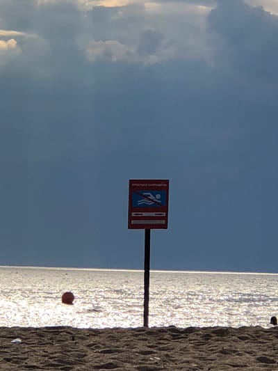 The image captures a beach scene in Sestroretsk, Russia, under a dramatic sky. The upper portion of the frame is dominated by heavy, dark blue-grey clouds, with some lighter patches indicating partial illumination. The horizon line is visible as a bright, thin strip where the dark sky meets the highly reflective sea.
The sea surface shimmers intensely with glints of light, creating a sparkling effect across the water. A single, distinct red spherical buoy floats in the water to the left of the image's center.
In the foreground, a dark sandy beach with visible undulations and footprints extends across the lower part of the frame. A small piece of white debris is discernible on the sand in the lower-left corner. On the far right edge of the beach, a dark, indistinct rounded shape, possibly a person's head or a small object, is partially visible.
Centrally positioned on the beach, a dark pole supports a rectangular red sign. The sign bears Cyrillic text at the top, which translates to "SWIMMING IS FORBIDDEN". Below the text, a graphic features a white swimmer symbol on a blue background, traversed by a red diagonal line, explicitly prohibiting swimming. Below this symbol, two white horizontal arrows point left and right on a blue background, likely indicating the boundaries of the forbidden area or directing people.