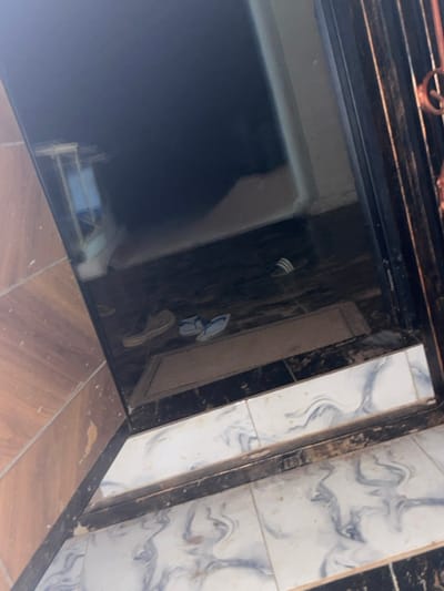 A corner of a room is visible with varying floor tiles and a shiny, reflective surface obscuring the lower left. There is a set of shoes and slippers scattered along the wall. A brown-toned wooden edge borders the tiles, and a wrought-iron railing runs along the top. The reflective surface shows the reflection of what appears to be a shelf with items on it. The lighting is bright, causing sharp contrasts in the scene.
