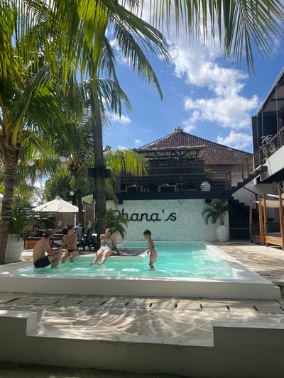 In Nusa Penida, Indonesia, a family enjoys a sunny day by a pool. A man with a beard and a maroon baseball cap, wearing black shorts, sits on the edge of the pool, holding a drink. Next to him, a woman in a black and white striped bikini is seated. To her right, another woman in a blue and white striped bikini reclines by the pool. A young boy, wearing orange patterned swim trunks, stands in the shallow end of the turquoise pool, with water up to his knees. Palm trees with lush green fronds overhang the scene, and in the background, a two-story building with a thatched roof and modern architecture is visible. White string lights are strung between palm trees, adding to the relaxed atmosphere. The ground around the pool is paved with irregular light-colored stones, and shadows from the palm trees create a dappled effect. The sky above is a bright blue with scattered white clouds.