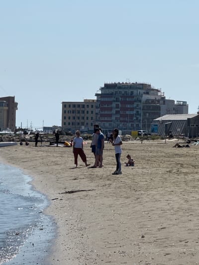 The image captures a beach scene in Anzio, Italy. Several mid-rise buildings form the backdrop against a clear sky. The foreground is dominated by the sandy beach where a small group of people stands. To the right, the edge of the water is visible. The group appears to be engaged in casual interaction. Further down the beach, other individuals are present, some reclining.