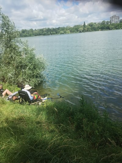 Two people are fishing from the shore of a body of water, likely a lake or river, in Sumy, Ukraine. The person in the foreground is sitting in a black fishing chair, wearing a wide-brimmed hat and colorful shorts, with fishing rods extending into the water. Another person is visible to the left, also seated and appearing to be fishing. The background shows a dense line of trees along the opposite bank, and beyond that, a city skyline with buildings. The sky is partly cloudy. The environment is natural and grassy, with a calm body of water.