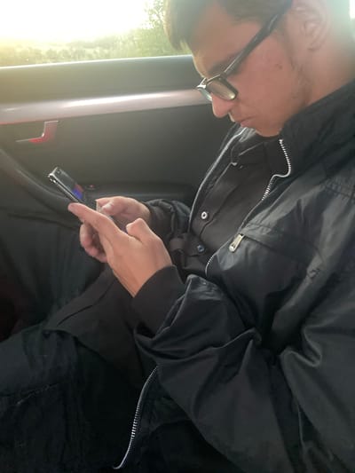 In the city of Ablanitsa, Bulgaria, a person wearing glasses and a black jacket is seated inside a vehicle, focused on their mobile phone. The individual is holding the phone in their hands, and their fingers are visible on the screen, suggesting they are actively using the device. The background shows the interior of the car, including the door handle and a glimpse of the outdoors through the window. The lighting suggests it might be daytime, possibly late afternoon due to the warm, bright light filtering in. The overall scene depicts a moment of personal activity within a vehicle.