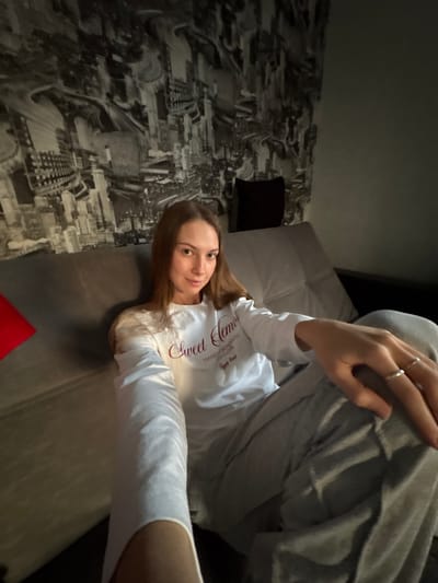 A young woman with long brown hair is sitting on a sofa in a dimly lit room. She is wearing a white long-sleeved shirt with text that appears to say "Sweet Dreams" and gray sweatpants. The background features a black and white mural of a city skyline. A red pillow is visible to the left of the woman. The overall scene suggests a casual and relaxed atmosphere. The location is not identifiable as Novaya Ivanovka, Russia based on the visual information provided.