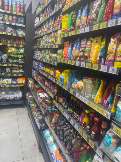 The image shows an aisle in a supermarket, likely in the city of Гожули, Ukraine, based on the context provided. The shelves are stocked with various food products, primarily packaged goods like sauces, condiments, snacks, and other grocery items. There are multiple shelves visible, each organized with rows of products. Some products are in plastic pouches, while others are in bottles or jars. Pricing labels are visible on some of the shelves. The lighting appears to be standard fluorescent supermarket lighting. The overall scene depicts a typical retail environment focused on food products.