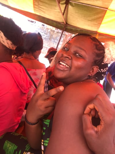 This image depicts a group of individuals in Akpaka Forest Reser, Nigeria, beneath an orange and olive green fabric canopy structure. In the foreground, a young adult female with dark skin and braided hair, interspersed with lighter strands, is shown smiling directly at the camera, revealing teeth. She wears small dark hoop earrings. Her left hand displays a V-sign, while her right hand forms an 'OK' gesture, resting on her right bicep. A dark bracelet is visible on her left wrist. Behind her and to the left, another individual is seen from the back, wearing a red top and a patterned headband. Further back, another person with a dark head covering and a light-colored patterned top is present. Additional individuals are partially visible in the background, including one wearing a red cap and another with light, curly hair. A person in a blue top with white lettering is partially visible on the far right. The background behind the individuals includes a light-colored, textured wall or structure. The scene is illuminated by natural light.
