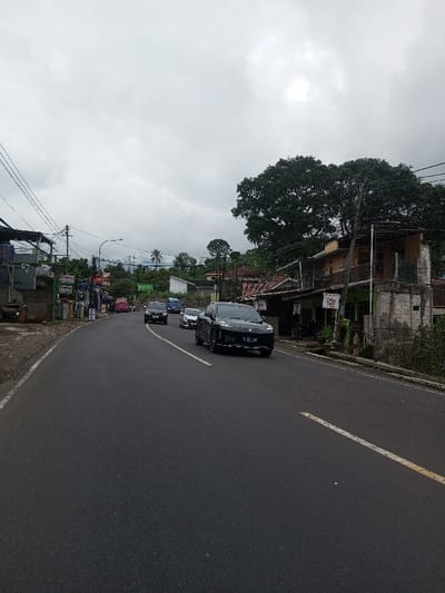 This image depicts a two-lane asphalt road curving to the left, situated in the city of Kodarapalli, India, under a heavily overcast sky. The road features a solid white line at its right edge and a broken yellow center line.
**Vehicles:**
A black SUV, bearing the license plate "B 2471 TMT," is prominent in the mid-foreground, moving towards the viewer on the right side of the road. It is followed by a silver sedan/hatchback, a dark-colored sedan/SUV, and a blue van/minibus, all traveling in the same direction. Further down the road on the left, a pink vehicle is visible.
**Right Side of the Road:**
A two-story building with a light brown upper facade and a light green lower section stands immediately adjacent to the road, featuring a small balcony on the upper level. Signs attached to this building include text that appears to read "WARUNG" and "AYAM GORENG." A large, dense green tree dominates the background behind this structure.
**Left Side of the Road:**
Multiple small structures, some with corrugated metal roofs, line the left side of the road. Utility poles with a dense network of overhead wires are visible. A blue container or water tank rests on a platform. Signs, including one displaying the word "Curuy," are affixed to these structures.
**Environment:**
The background features rolling hills covered in lush green vegetation and trees, including some palm trees. The sky is completely covered by thick, grey clouds, indicating an overcast condition with diffused light. No individuals are distinctly visible in the scene.