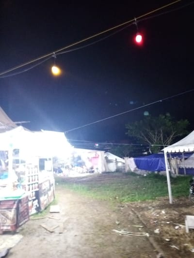 This nighttime photograph captures an outdoor scene, likely a temporary market or gathering, possibly within Khe Sanh Commune, Vietnam. The dark sky dominates the upper portion of the image, punctuated by artificial lights.
In the foreground on the left, a brightly illuminated stall with a white canopy stands out. The intensity of its light source creates an overexposed effect, obscuring precise details within the stall, though rows of bottles or jars can be faintly seen, suggesting it might be a beverage or food vendor. The ground in front of this stall is a mix of dirt and mud, with some debris visible.
A dirt path or open area extends from the foreground into the mid-ground, leading toward several other temporary structures. These include various tents and makeshift shelters, some covered with white tarpaulins and others with blue tarps, indicating an informal or temporary setup. In the background on the right, dark tree foliage is visible against the night sky. On the far right, part of a white tent and a section of a bicycle wheel can be observed.
Above, two string lights are suspended across the dark expanse. One light on the left glows with a warm yellow-orange hue, while another further to the right emits a distinct red light. The overall illumination is sparse, with the prominent stall on the left providing the most intense light. No individuals are visible in the photograph, lending a quiet atmosphere to what appears to be a communal space or event. There is no discernible text present within the image.