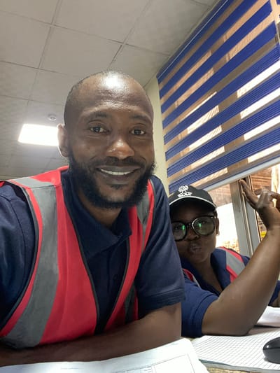 This portrait-oriented image, rotated 90 degrees clockwise, captures two individuals in what appears to be an indoor work environment in Sabon Gari Nassarawa, Nigeria.
In the foreground, a man with dark skin, a neatly trimmed beard, and short hair is smiling broadly, looking directly at the camera. He is wearing a navy blue collared shirt beneath a red and grey reflective safety vest.
Partially visible behind him is another individual with dark skin, wearing a similar navy blue shirt. This person is also wearing a black baseball cap with an indiscernible white emblem and round-rimmed glasses. Their left arm is raised, hand open and palm facing upwards, suggesting a gesture or pointing towards something. They appear to be looking upwards or away from the camera, with a slightly pursed expression.
The background features bright overhead fluorescent lighting, with a distinctive ceiling or high window system composed of alternating blue and light-colored slats or blinds. To the right of the partially obscured person, several white documents or papers are visible on a surface. Faint text on these papers includes phrases like "PLEASE" and "Payment Terms," indicating a professional or administrative context. The matching shirts and reflective vests suggest that both individuals are colleagues, likely employees in a role where visibility and uniform are required, such as logistics, industrial, or service work. The scene conveys a brightly lit work environment.