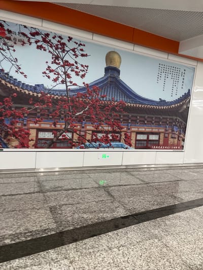 The image showcases a vibrant mural depicting a traditional Chinese building with a distinctive blue, curved roof and a golden dome. Red blossoms from a flowering tree in the foreground add a striking splash of color against the building's architectural details. The mural is installed on a wall, possibly in a public space given the presence of what appears to be a subway station setting with tiled flooring and directional signage.
The mural itself features text in Chinese calligraphy, with some phrases relating to the cotton tree ("木棉") and its symbolism. The text, when translated, speaks of the tree's resilience and its connection to the spirit of the people of Guangzhou (referred to as "羊城人" or "Goat City people"), suggesting it as a symbol of pioneering spirit. There is also a phone number displayed, likely for information or services related to the mural or the location.
The lighting suggests it is daytime, and the overall mood is one of cultural appreciation and artistic display. The juxtaposition of the traditional artwork with the modern, clean lines of the surrounding environment implies a contemporary urban setting. The tiled floor and the metallic accents on the wall further reinforce the idea of a well-maintained public space. The presence of green illuminated signs with arrows suggests a transit hub or a similar public facility. The overall impression is a blend of cultural heritage and modern infrastructure.