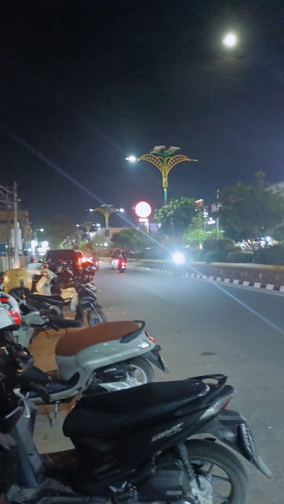 This media file captures a nighttime scene along a street in Bireuen, Indonesia. The foreground is dominated by several parked motorcycles, with their seats and parts of their bodies visible. The closest motorcycle has a black seat and fender, while another to its left features a brown seat and a lighter-colored body. Helmets are also visible near the parked scooters.
The street is lit by streetlights, some of which have distinctive decorative designs resembling stylized trees with radiating yellow elements. A prominent red circular sign is visible in the mid-ground. The road itself is visible, with a white and black striped barrier along the side. Vehicles, including what appears to be a van and several motorcycles, are moving along the street, their headlights and taillights illuminating the darkness.
In the sky, a bright full moon is clearly visible, casting a gentle light. The overall atmosphere is one of urban nighttime activity, with the combination of artificial lighting from the streetlights and the natural light from the moon creating a serene yet active ambiance. The presence of numerous motorcycles suggests a common mode of transportation in this location. The scene appears calm, with no direct indication of any specific event beyond ordinary street life at night. No discernible text is visible in the image.