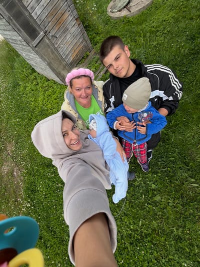 In the city of Andreyevo, Russia, a group of four individuals is captured in a high-angle selfie taken outdoors. The setting is a grassy area, possibly a yard or park, with a wooden structure visible in the upper left corner, resembling a shed or outbuilding.
The individuals appear to be two women, a teenage boy, and two young children. One woman, wearing a light beige hooded sweatshirt, holds a baby wrapped in a light blue swaddle. She is smiling directly at the camera. Another woman, positioned behind her, wears a green shirt and a pink padded headband. She is also smiling. The teenage boy, dressed in a black and white Adidas jacket, is holding a toddler. The toddler is wearing a blue jacket and red plaid pants, with a light grey beanie on his head. His tongue is sticking out slightly. The scene suggests a family gathering or outing.
