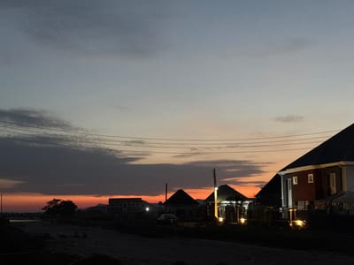 This image captures a dusk scene in Angwan Sayawa, Nigeria. The sky is characterized by a gradient from light blue-grey at the zenith, transitioning to a band of darker grey clouds, and culminating in a vibrant orange-red glow along the horizon, signifying sunset. Multiple parallel power lines traverse the upper-mid section of the frame.

The foreground is composed of dark, unpaved ground, suggesting an undeveloped road or construction area, with visible rough terrain. No human figures are present.

On the left, a dark tree is silhouetted against the orange sky. Distant, dark structures, likely buildings, are faintly discernible, and a faint light source, possibly a street lamp, illuminates a small patch of ground.

The mid-ground and right side of the image feature several residential buildings. A dark sedan car is parked on the unpaved surface. Behind it, a house with a dark, peaked roof is largely silhouetted. To the right, another house with a green-tiled roof exhibits illuminated exterior lighting. A prominent two-story house occupies the right foreground, featuring a dark, sloped roof, a reddish-brown exterior section with white-framed windows, and multiple exterior lights casting warm yellow illumination on its facade and the adjacent dark ground, where fencing and sparse vegetation are visible. Utility poles with overhead wires are interspersed among the structures. In the far distance, low mountains or hills are visible as dark silhouettes against the horizon.