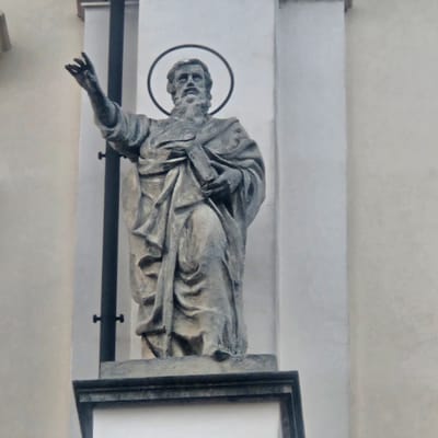 The image shows a stone statue of a bearded man with a halo, holding a book. The statue is mounted on the exterior of a building. The man is depicted with his right arm raised and his hand open, as if in greeting or blessing. His left arm is bent, and he holds a book or scroll against his chest. The statue is made of weathered stone, with visible details in the drapery of his robe and his facial features. The background is a plain, light-colored wall, possibly stucco or concrete. A dark vertical pole is visible to the left of the statue. The statue is positioned above a dark, rectangular base.

This statue is located in Calcinate, Italy, specifically on the façade of the Church of San Pietro.