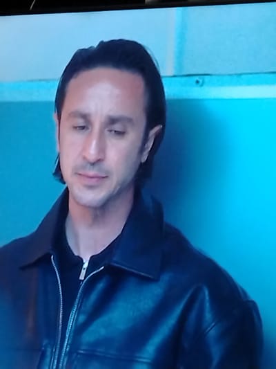 The image is a medium shot of a man with dark hair, wearing a black leather jacket over a black zip-up shirt. He is looking down with a neutral expression. The background is a light blue wall with a horizontal white stripe near the top. There is no indication of location.