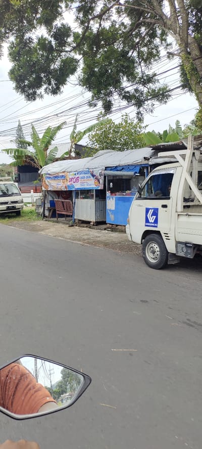 The image shows a street scene in Kecamatan Lawang, Indonesia (latitude: -7.852, longitude: 112.692). A white pickup truck is parked on the right side of the road. The truck has a blue logo with the words "Langgeng H/L" on the side. The truck is facing the camera. It is parked in front of a small store with a white roof and blue walls. The store has a sign with a logo and the words "PROMO 1025-2025 TENDA BIRU" written on it. There are power lines running overhead and a large tree to the left of the truck. A white van is parked further down the road on the left side. The asphalt road is smooth and gray. The mirror of a motorcycle is reflecting a scene of trees, buildings, and overhead power lines. The sun is shining. The overall environment is calm and mundane.