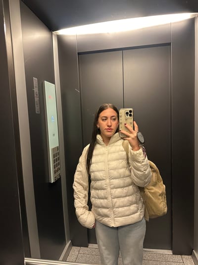 A woman is taking a selfie in an elevator. She is wearing a white puffer jacket and grey sweatpants, and she has a tan corduroy bag slung over her shoulder. She holds a cell phone in her right hand. The elevator has a gray interior with a horizontal button panel. There is a strip of bright white light on the left side of the elevator. The location is presumed to be somewhere in the city of Huixquilucan de Degollado, Mexico, based on the user's request.