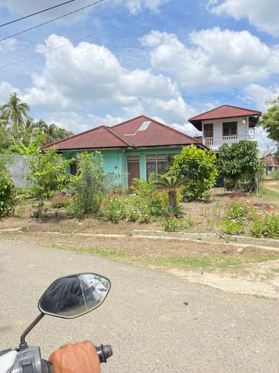 A residential scene in Langsa, Indonesia, is depicted from the perspective of someone riding a motorbike. The foreground features the handlebars and a portion of the motorbike's side mirror, reflecting a dark object and a white patterned fabric. The road surface is asphalt.

The midground showcases a modest house with a turquoise-painted facade and a reddish-brown corrugated metal roof. Adjacent to the main house is a two-story structure with a white upper level featuring a balcony, also topped with a reddish-brown roof. The house is surrounded by a garden filled with various flowering plants in shades of pink, white, and red, along with lush green foliage and trees. Palm trees and other tropical vegetation are visible in the background, suggesting a warm climate.

The sky is a clear blue with scattered fluffy white clouds, indicating fair weather. The overall impression is of a rural or suburban residential area in Indonesia.