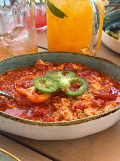 A close-up, eye-level shot features a rustic bowl of a red, chunky stew, possibly shakshuka, garnished with bright green jalapeño and orange bell pepper slices. The stew fills the foreground, with a silver spoon resting on the left edge of the bowl. In the background, out of focus, are two tall glasses filled with amber and pink beverages, garnished with mint and lime, suggesting a restaurant or cafe setting. The wooden tabletop surfaces the setting. The image is taken at "Aghasi" restaurant in Byurakan, Armenia, a location known for its celestial views and tranquil atmosphere.