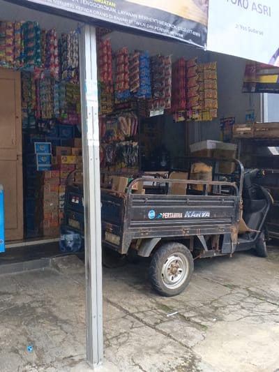 The image depicts the front exterior of a small shop, or *warung*, in Ciamis, Indonesia, during daylight hours. A black, three-wheeled cargo motorbike is parked directly in front of the shop entrance.

The shop's entryway is heavily adorned with numerous sachets of snacks or instant foods, hung in vertical displays. Inside, stacks of cardboard boxes, likely containing bottled drinks or other provisions, are visible. The ground in front of the shop is concrete, showing some cracks and minor discoloration. A silver-colored support pillar stands prominently in the foreground, off-center.

A white banner spans the top of the shop entrance. Visible text on the banner includes "ASRI" (possibly part of the shop's name), "Jl Yos Sudarso" (a street name), and Indonesian text that appears to be a public health warning: "MEMBERI KEPADA ORANG DI BAWAH 21 TAHUN DAN PEREMPUAN HAML" (giving/selling to people under 21 and pregnant women) and "LAYANAN BERHENTI MEROKOK (0800-177-65051" (quit smoking service helpline).

The cargo motorbike has "PERTAMINA KARYA" written on its side, accompanied by a logo, indicating a connection to Pertamina, the Indonesian state-owned oil and gas company. Its cargo bed holds a few cardboard boxes. No individuals are visible in the scene. The image captures a common commercial street-side tableau in Indonesia.