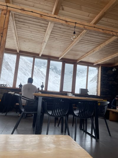 This image captures the interior of a cafe or restaurant situated in Juta, Georgia, offering a panoramic view of a snow-covered mountain valley. The establishment features a ceiling constructed from light-toned wooden planks supported by exposed wooden beams, from which bare spherical light bulbs are suspended via black wiring. The floor is a dark grey concrete. The far wall is dominated by a series of large, vertical-paneled windows, framed in wood, showcasing a vista of steep, snow-covered mountains beneath an overcast sky.

The interior is furnished with multiple black, modern-style plastic chairs featuring slatted backs and seats, arranged around tables with dark frames and light wooden tops. A long wooden counter runs beneath the windows, displaying decorative objects including two dark-colored pitchers or vases and a smaller dark figurine. A partially visible "CAFE" sign is affixed to the counter's left side. A rectangular dark grey object, possibly an appliance, rests on the counter to the right.

Two individuals are present. A male subject is seated centrally, viewed from the back, wearing a light-colored (possibly white or light grey) short-sleeved shirt, with a distinct dark stain visible on the upper left back. He is positioned facing the windows, observing the outdoor landscape. A second male subject is partially visible on the far right, seated at a separate wooden table against a section of dark, stacked stone wall. He wears a dark cap and dark attire, and appears to be interacting with an object held near his mouth.