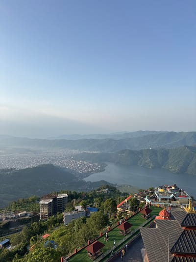 The image is an outdoor daytime scene featuring a mountain range, a lake, a city, and a structure on a rooftop with people. The sky is clear and blue. The mountain range stretches across the middle of the frame, partially obscured by haze. A large lake winds through the mountains. To the right of the lake, a city is nestled in the valley. The foreground features a rooftop structure with a red-tiled roof and a green patio area with several small gazebos. People are present on the patio, some standing and some seated. The location is likely Pokhara, Nepal, based on the landscape and architecture.