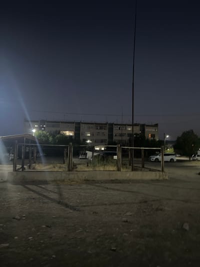 A long shot image taken at dusk shows a multi-story apartment building in the city of Şamaldı-Say, Kyrgyzstan. The building has many windows, some of which are illuminated, suggesting occupancy. In front of the building, there are several trees and a wooden structure that appears to be a shelter or a part of a playground. The foreground of the image is a dirt road with scattered rocks and debris. A few cars are visible in the background, parked on the side of the road or in a lot. A tall pole stands near the apartment building, and a street light is illuminated in the distance. The overall scene depicts a residential area at the end of the day.