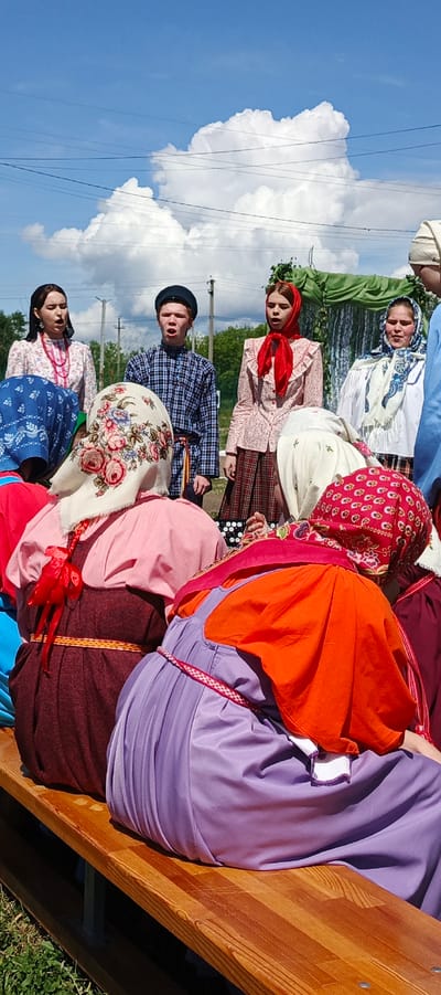 The photograph depicts a group of people gathered in what appears to be a performance setting in the city of Кемуль, Russia. In the foreground, several people are seated on a wooden bench, their backs to the camera. They wear traditional Russian clothing, including brightly colored dresses and headscarves with floral patterns.
Behind the seated individuals, a small group of people stands and faces the camera. They also wear traditional Russian clothing. The backdrop includes trees, power lines, and a large white cloud in a blue sky. The overall setting is outdoors, and the photograph appears to have been taken during a sunny day.