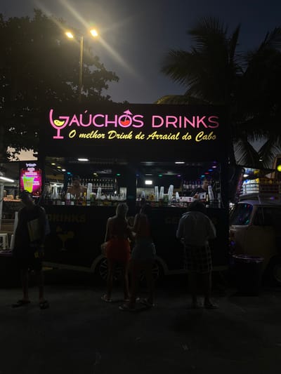 A lit-up "GAÚCHOS DRINKS" sign with pink accents stands beside a dark road at night. The sign boasts "O melhor Drink de Arraial do Cabo." A mobile bar draws a small crowd. Customers cluster around ordering drinks from the bartender. A few pedestrians pass by. A palm tree looms in the background near a street light, partially obscuring the darkening sky.