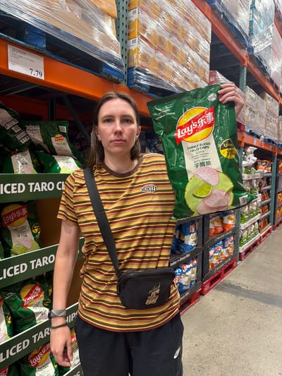 In Beaverton, Oregon, a woman stands in a Costco store, holding a large bag of Lay's thinly sliced taro chips with lime flavor. She is wearing a horizontally striped t-shirt in shades of brown and yellow, black shorts, and a black fanny pack with a graphic detail. She has on a watch and a bracelet on her left wrist. In the background, shelves are stocked with various chips and food products. The store environment appears to be a typical warehouse-style retail setting.