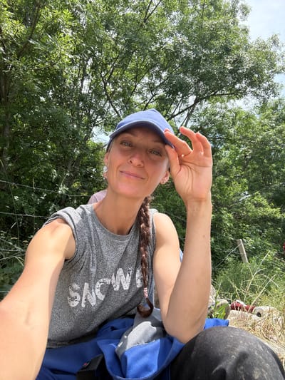 A woman with a braided brown ponytail and a blue baseball cap is taking a selfie outdoors. She is wearing a grey t-shirt with the word "SNOW" printed on it in white letters. Her right arm is bent at the elbow and her fingers are touching the brim of her cap. Her left arm is also bent at the elbow and resting on her lap. She is seated on a blue and grey bag, and black pants are visible. In the background, there are green trees and a wire fence. A blurred figure in a pink shirt is partially visible behind her. The scene appears to be in a rural or natural setting, possibly on the outskirts of Lavrovoe, Ukraine, given the presence of dense vegetation and a rustic fence. The lighting suggests it is a sunny day.