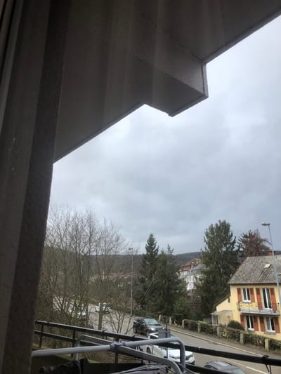 The image depicts an exterior view from an elevated position, likely a balcony or window, overlooking a residential street in Saverne, France, under an overcast sky. In the foreground, the underside of a textured, grey concrete structure, possibly a balcony or eaves, spans the top and upper left, with a vertical concrete pillar or wall section on the far left. A metallic clothes drying rack with dark fabric is partially visible at the bottom center of the frame.

The midground features a paved road curving from left to right. On this road, a dark-colored SUV, possibly black or dark grey, is centrally positioned with its headlights illuminated, facing towards the right. A white car, possibly a Renault Clio V, is parked behind the SUV, partially obscured. Another dark-colored vehicle is visible further down the road to the right, positioned beyond a yellow building. The roadside is lined with bare deciduous trees and several evergreen conifers. Streetlights are visible, one prominent on the right side of the road, another further back on the left. Low stone walls topped with hedges are present along the road, particularly in front of the yellow building.

In the background, a multi-story yellow building with multiple windows and reddish-brown shutters is prominent on the right. This building features an ornate metallic balcony railing on an upper floor and has a grey sloped roof. Further back and to the left of the yellow building, other residential structures with varying architectural styles and roof colors are visible amidst the trees. Distant hills, largely covered with bare trees, form the horizon. The sky is uniformly light grey and overcast, indicating a lack of direct sunlight. No individuals are visible, and no discernible actions or interactions are depicted.