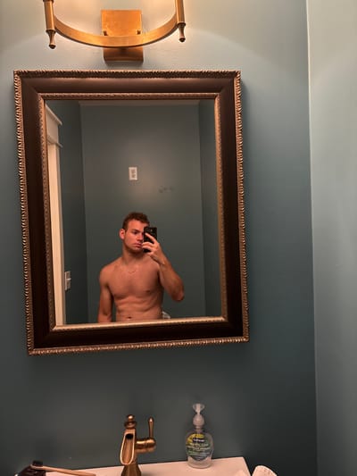 A shirtless man is taking a selfie in a bathroom mirror. He is standing in front of a teal-colored wall and a large, ornate mirror. The mirror reflects a light fixture above it, an electrical outlet on the wall, and a doorway on the left. The man is holding a black smartphone in his right hand. Below the mirror, a gold-colored faucet is visible, along with a bottle of hand soap and a toothbrush. The overall scene suggests a residential bathroom. The location is not specified in the image or the provided crops.