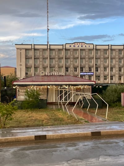 The image depicts the Hotel Xojeli and a ground-level building with a covered outdoor seating area in Khujayli, Uzbekistan. The hotel is a multi-story building with a concrete facade decorated with geometric patterns and a central sign reading "HOTEL XOJELI". Below the hotel, a red-roofed structure houses a dining or cafe establishment, indicated by a sign that appears to say "BAUYSAO" and "BREND CINEMA". String lights are draped across the outdoor seating area, which features white hexagonal metal frames leading to the entrance. The foreground shows a wet paved area, likely a street or sidewalk, with a concrete curb. The environment appears to be overcast, with a tall communication tower visible in the background. There are no people visible in the image.