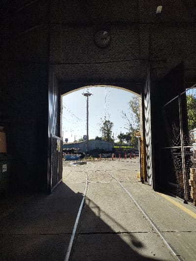 The image depicts a view looking out from inside a darkened, brick-lined arched gateway into a brightly lit industrial or railway yard in Sáenz Peña, Argentina. In the foreground, two parallel railway tracks are embedded within a concrete floor, extending from the shadows of the interior outward. To the immediate left, a dark vertical structure partially obscures a green industrial bin topped with a brown cardboard box. To the right, another dark vertical structure, likely a large door or gate, features a metal grid panel, with a yellow stripe visible on the adjacent concrete floor. A circular clock face is mounted on the brickwork above the archway, its details indistinct.
Beyond the archway, the exterior ground is a mix of concrete and possibly dirt, illuminated by direct sunlight. A series of orange traffic cones are arranged in a line in the midground. A prominent tall, light-colored mast with a circular platform at its top stands against a clear, light blue sky in the center-left. Faint lines, possibly overhead wires, crisscross the sky. Behind the mast, a low-slung building constructed of light blue corrugated metal is visible. To the right of the tracks in the mid-distance, a small yellow rectangular sign is affixed to a pole. Green trees are visible further in the background to the right. No individuals are present in the depicted scene.