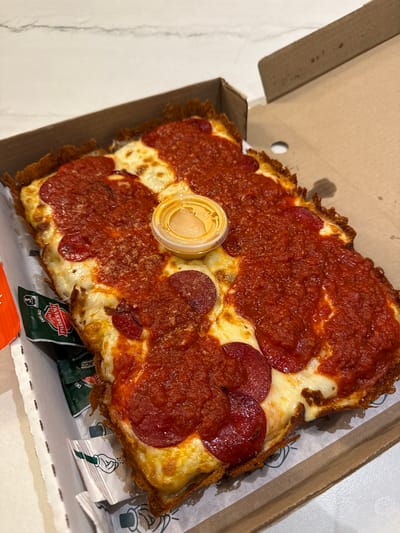 A partially consumed pepperoni pizza is in a cardboard box. A small, clear plastic container with a yellow sauce sits on top of the pizza. Also in the box are several packets of Parmesan cheese and pepper. The pizza has a thick, crispy crust. Based on the logo on the Parmesan packets, the pizza is from Peter Piper Pizza. The packaging says that the Parmesan is provided by DEL AGG, a Mexican food supplier. Peter Piper Pizza has a location in Monterrey, Mexico. Therefore, the image is likely from there.