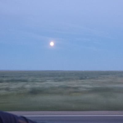 The image shows a wide, flat landscape under a pale blue sky with a full moon visible. The foreground features a section of a dark road with a white line. The middle ground is dominated by a blur of green and grey, suggesting fields or vegetation that are out of focus due to motion. The horizon line is low and indistinct, where the landscape meets the sky. The scene appears to be captured from a moving vehicle, indicated by the blur in the middle ground. The location is identified as Aktobe, Kazakhstan. There are no people or specific objects clearly discernible in the landscape, with the exception of the road and the moon. The overall impression is one of open, vast space and a sense of travel.