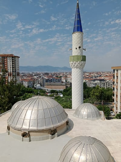A high-angle, medium shot captures a mosque's domes and minaret against a backdrop of a bustling cityscape under a bright, partially cloudy sky. The foreground features three silver domes with segmented, reflective surfaces, two of which are large and prominent, and a third, smaller dome positioned to the right. The domes sit atop a white roof. To the left, a tall, white minaret rises, adorned with a textured pattern and topped with a striking blue, faceted spire. Two speaker horns are visible on either side of the minaret's midsection, and a green band encircles its base.
Behind the mosque structures, the urban landscape of Bayraklı, Turkey, unfolds. Numerous buildings with red-tiled roofs and varying architectural styles fill the middle ground, interspersed with lush green trees. A large, modern apartment building with a brown and white facade stands prominently to the left. In the distance, hazy mountains frame the horizon. The sky is a vibrant blue, dotted with wispy white clouds, suggesting a clear, sunny day, likely in the morning or afternoon given the bright light and sharp shadows. No people are visible in the image, and there is no discernible text. The scene conveys a sense of urban life integrated with religious architecture.