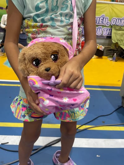 In Thuận An, Vietnam, a young girl is captured in a close-up shot, holding a plush toy dog. She is wearing a light blue t-shirt with a colorful floral print and a brightly patterned skirt. The plush dog, with large, expressive eyes and a brown furry body, is wrapped in a pink blanket adorned with rainbows, hearts, and flowers. The girl's hands are visible, gently holding the toy. The background suggests an indoor environment with a bright yellow floor marked with blue and white lines, possibly a gymnasium or recreational area. A partial view of a banner with Vietnamese text is also visible in the background. The overall scene conveys a sense of childhood playfulness and affection.