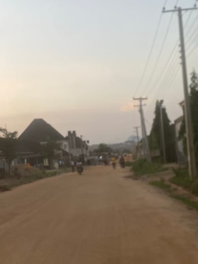 This blurry wide-angle shot depicts an unpaved road scene in Lugbe, Nigeria, during twilight conditions, characterized by a gradient sky transitioning from pale blue-grey overhead to warm yellow-orange near the horizon. The foreground and middle ground feature a wide, light brown dirt road, showing signs of vehicle and pedestrian traffic.

Along the left side of the road, several buildings are visible, including a prominent structure with a dark, steeply pitched roof resembling a pyramid. Adjacent to it are other multi-story buildings with lighter facades. Sparse green foliage, including trees and bushes, is interspersed among these structures. A few indistinct human figures are present near the buildings, and one individual on a motorcycle is captured in motion on the left side of the road, heading away from the viewer.

The right side of the road is lined with multiple utility poles carrying numerous electrical or communication wires, extending into the distance. Tall, slender, dark green trees stand behind the initial utility poles, partially obscuring additional structures further down the road. Another, smaller utility pole is visible further into the mid-ground on the left of the main pole series.

In the mid-ground, on the dirt road, at least two blurred human figures appear to be walking, one distinguishable by a yellow top. The far background reveals distant hills or elevated terrain under the soft, diffused light of the setting or rising sun. The overall scene suggests a semi-urban or developing area with essential infrastructure.