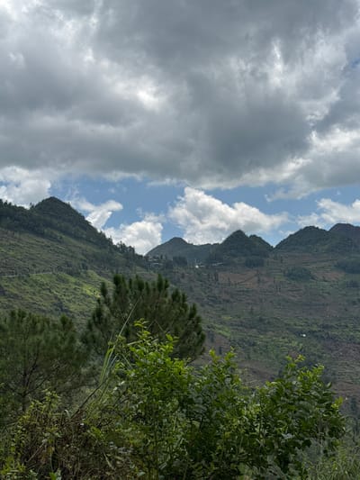 The image shows a mountain landscape in Lũng Cú Commune, Vietnam. The mountains are covered in green vegetation with some terraced fields visible. The sky is partly cloudy with both dark storm clouds and patches of blue. The right side of the image is filled with dense trees and foliage.