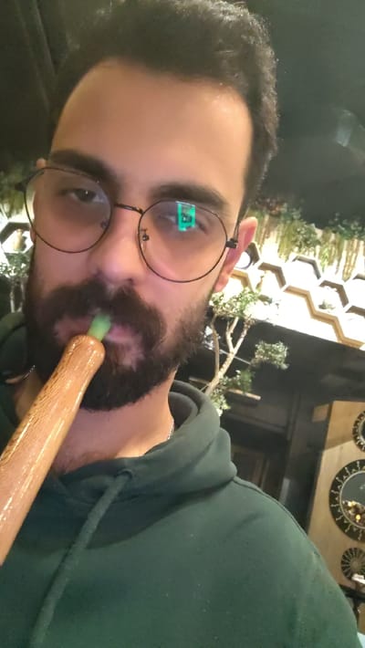 A selfie shows a man with a beard, dark hair, and glasses wearing a green hooded sweatshirt. He is holding a hookah pipe to his mouth, inhaling or exhaling. The reflection in his glasses reveals a blurred image, potentially a window or light source. The background is a mix of light-colored decorative elements and darker structures. The man is positioned in the left foreground, while the background is more distant and less focused.