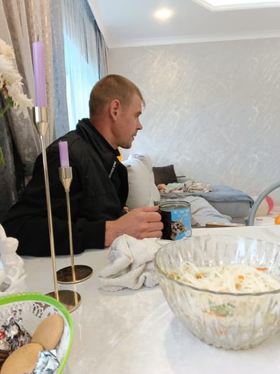 A photograph shows a man seated at a table inside a room. The room is identified as being located in Andreyevo, Russia. The man is fair-skinned with short, light-colored hair, wearing a black jacket. He is holding a mug with a cartoon cat design and the text "ALL YOU NEED IS LOVE". On the table in front of him is a bowl of what appears to be salad, a white towel, a decorative Easter basket with cookies and wrapped sweets, and two gold candlesticks holding purple candles. In the background, a person appears to be lying on a gray couch or daybed. The walls of the room are light-colored with a textured pattern. A curtain hangs near the side of the frame. A single light fixture is centered on the ceiling.
