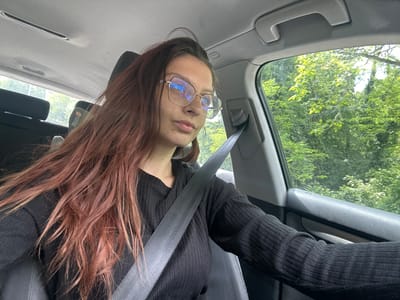 A young woman with long, reddish-brown hair is sitting in the passenger seat of a car. She is wearing a black, ribbed, long-sleeved shirt and large, gold-rimmed glasses. Her seatbelt is fastened across her chest. She is looking slightly to her right, with a neutral expression. The car interior is visible, including the dashboard, headliner, and a portion of the car door. Outside the car window, lush green trees and foliage are visible, suggesting a rural or suburban setting. The scene appears to be captured during daylight hours, with natural light illuminating the interior. The location is identified as Nikolaevo, Bulgaria.