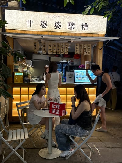 The image shows an outdoor storefront at night, likely in the city of Nan'an District, China, given the Chinese characters on the illuminated sign that reads "甘婆婆醪糟", translated as "Gan Popo Fermented Rice Wine" and indicating a food establishment specializing in a traditional Chinese beverage. Other smaller signs listing prices, and possibly menu items, flank the central sign. Four people are visible: a man studying a digital menu display, a woman looking at a digital phone, and two women seated at a round table eating or drinking. The storefront has an orange-toned grid pattern on the wall behind the counter and menu. Folding metal chairs surround the tables. The scene is dimly lit, with some greenery present near the bottom left of the frame. A red sign displaying Chinese characters is visible on the table.
