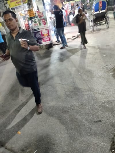 The image shows a man walking on a city street in Dhaka, Bangladesh. The street is paved with grey concrete. The man is wearing a grey shirt and blue jeans. He is holding a cup in his right hand and is walking away from the camera. There is a shop with a sign that reads "Indian Corner" in the background. Several other people are visible in the distance, and a blue rickshaw is parked on the side of the street. The environment is urban, with buildings on either side of the street. The coordinates provided indicate the location is in the densely populated neighborhood of Dhanmondi.