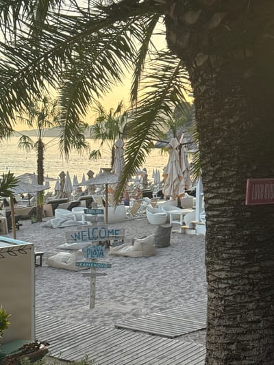 The image captures a sandy beach scene in Kamenovo, Montenegro, likely at dusk given the warm, golden light. In the foreground, the textured trunk of a palm tree dominates the right side of the frame, with its fronds partially obscuring the background. To the left of the palm tree, wooden planks form a pathway across the sand.
The beach itself is dotted with lounge chairs and umbrellas, many of which are closed and stacked. Several white, modern-style seating arrangements, including sofas and tables, are visible. A few beanbag chairs are scattered on the sand.
A wooden signpost stands in the middle ground, featuring two signs. The upper sign reads "WELCOME" in blue lettering, and the lower sign has "PLAYA" and "KAMENOVO" written on it, with an arrow pointing to the right. Further back, the calm sea stretches to the horizon, with a distant shoreline visible. A few people are sparsely scattered on the beach, some seated in lounge chairs. A pink sign with the text "LOVE EV..." is affixed to the palm tree trunk.