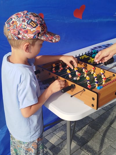 A young boy wearing a colorful patterned baseball cap and a light blue t-shirt plays foosball at a white table set up outdoors. He is looking intently at the game. Another person's hands are visible on the right side of the foosball table, also playing the game. The background is a solid blue fabric, with two red heart cutouts attached to it, one large and one small. The table has a wooden finish and a green playing field with miniature players. The ground is paved with grey interlocking stones. The scene appears to be an outdoor event or fair. The location is the city of Zavodoukovsk, Russia.